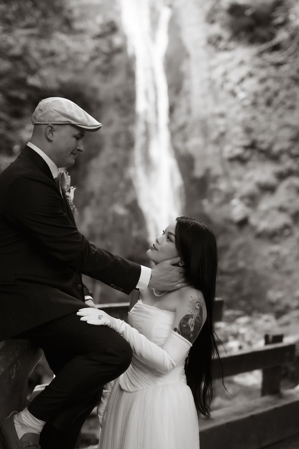 Black and white wedding photo of bride and groom sharing romantic moment in front of waterfall.