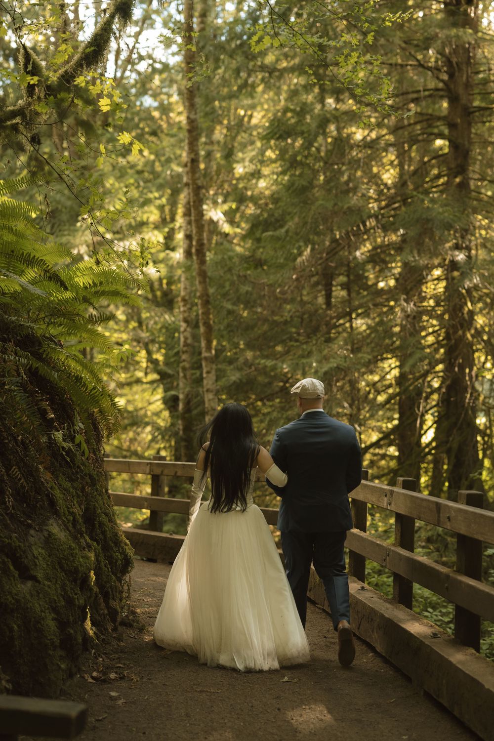 Bride and groom walking hand in hand along a wooden path through a lush green forest.