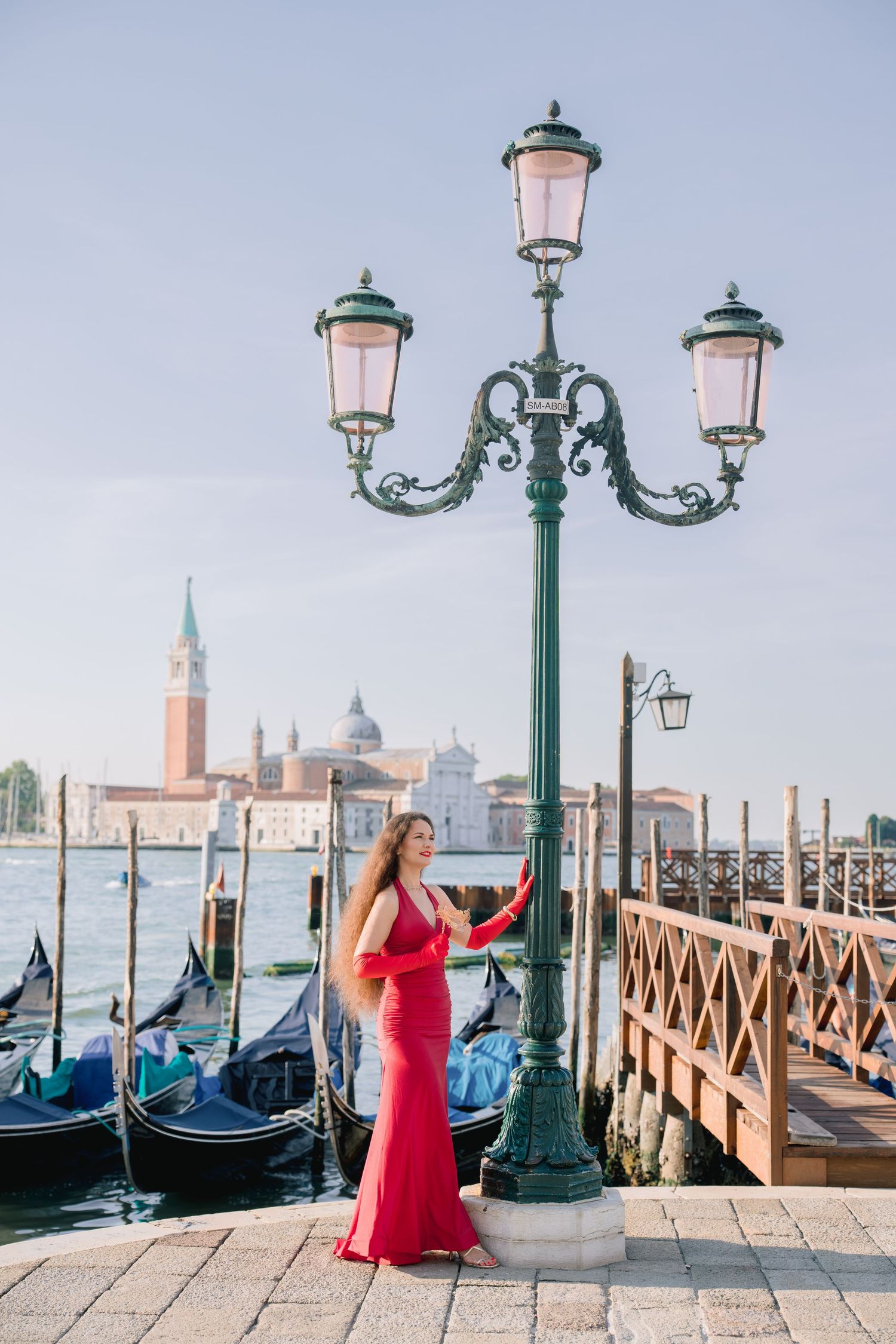Woman in red gown poses by ornate lamppost with Venice gondolas and San Giorgio Maggiore island in background.