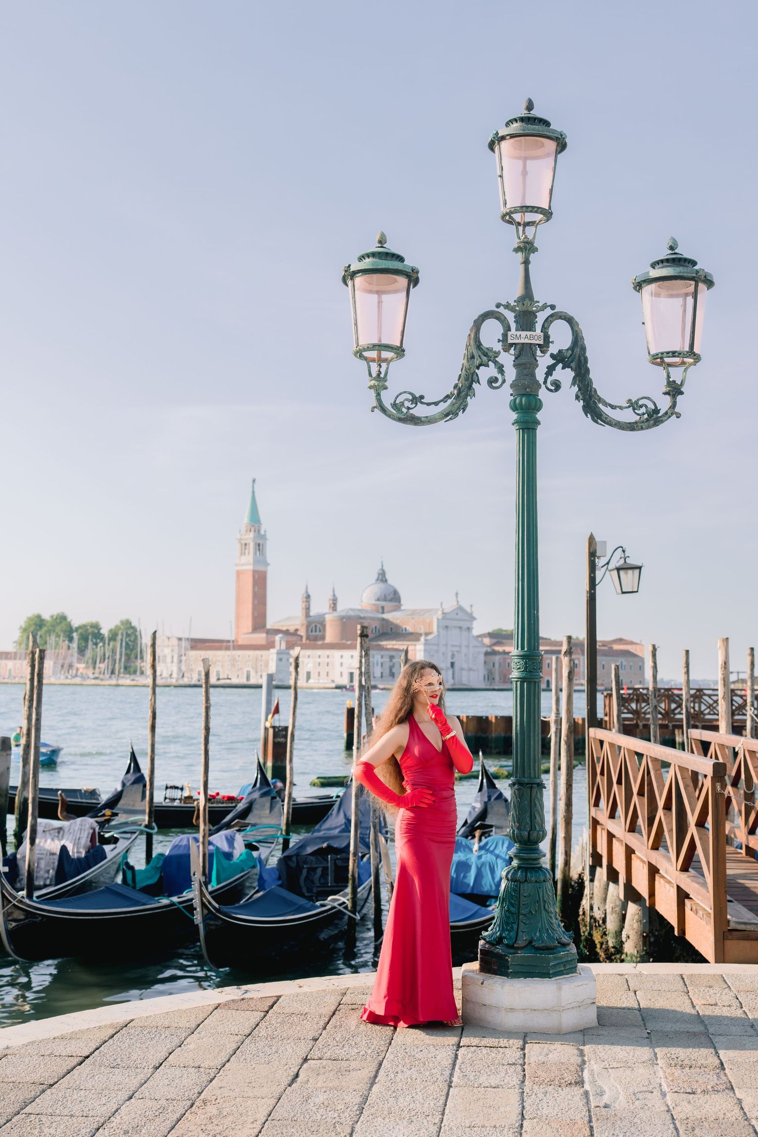 Woman in red gown poses by ornate lamppost with Venice gondolas and San Giorgio Maggiore island in background.