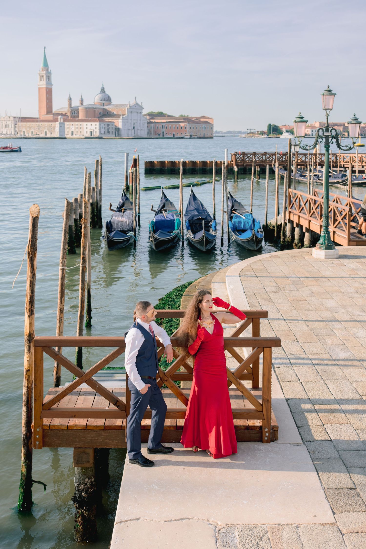 Couple in Venice by gondolas, woman in red dress and man in blue suit, with San Giorgio Maggiore in background.