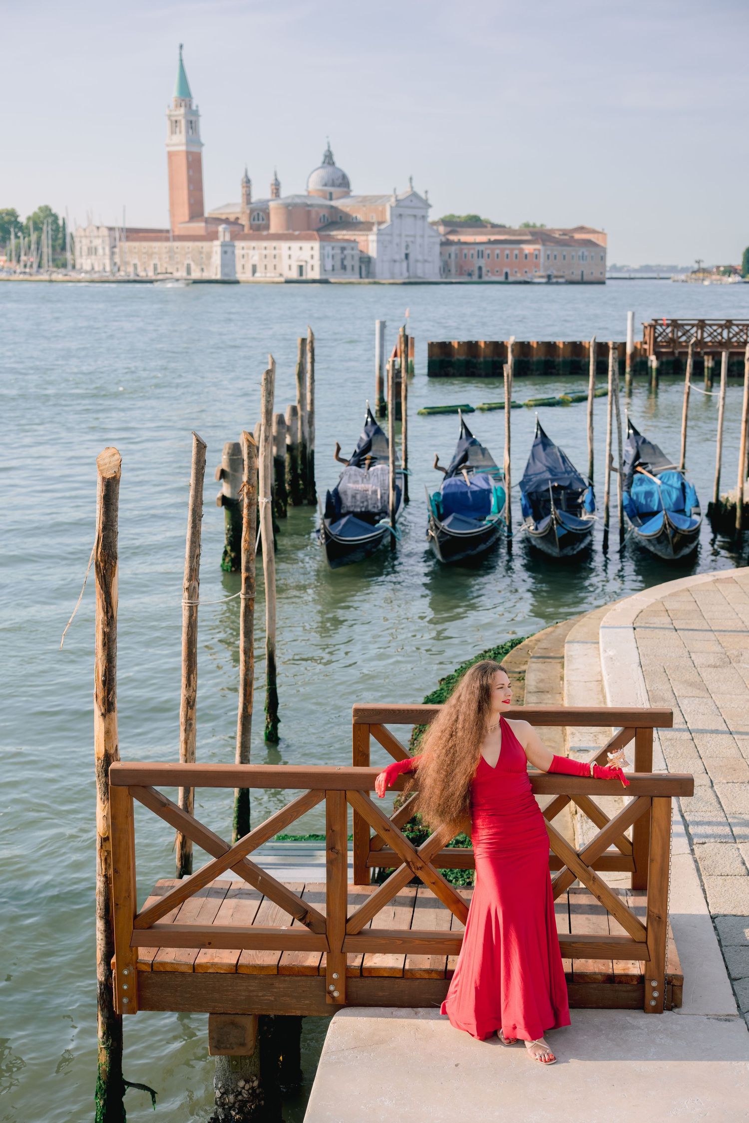 Woman in red dress poses on Venice waterfront dock with gondolas and San Giorgio Maggiore island in background.