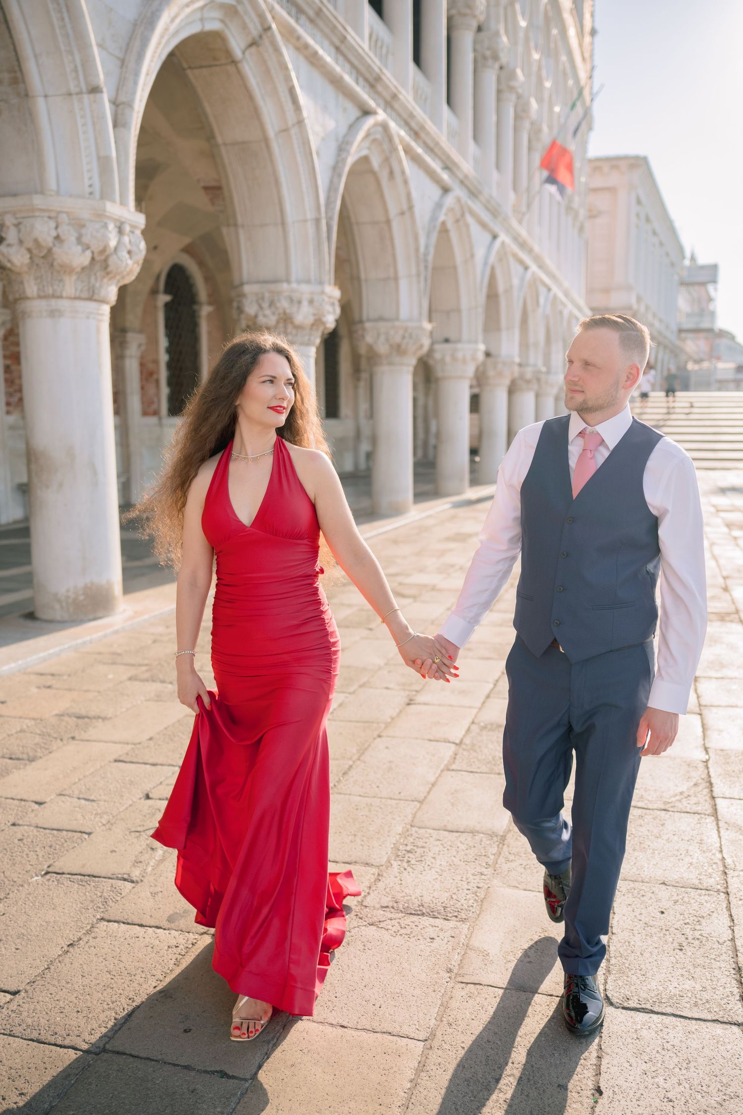 Couple in formal wear holding hands at Venice's Doge's Palace, woman in red gown, man in navy suit.