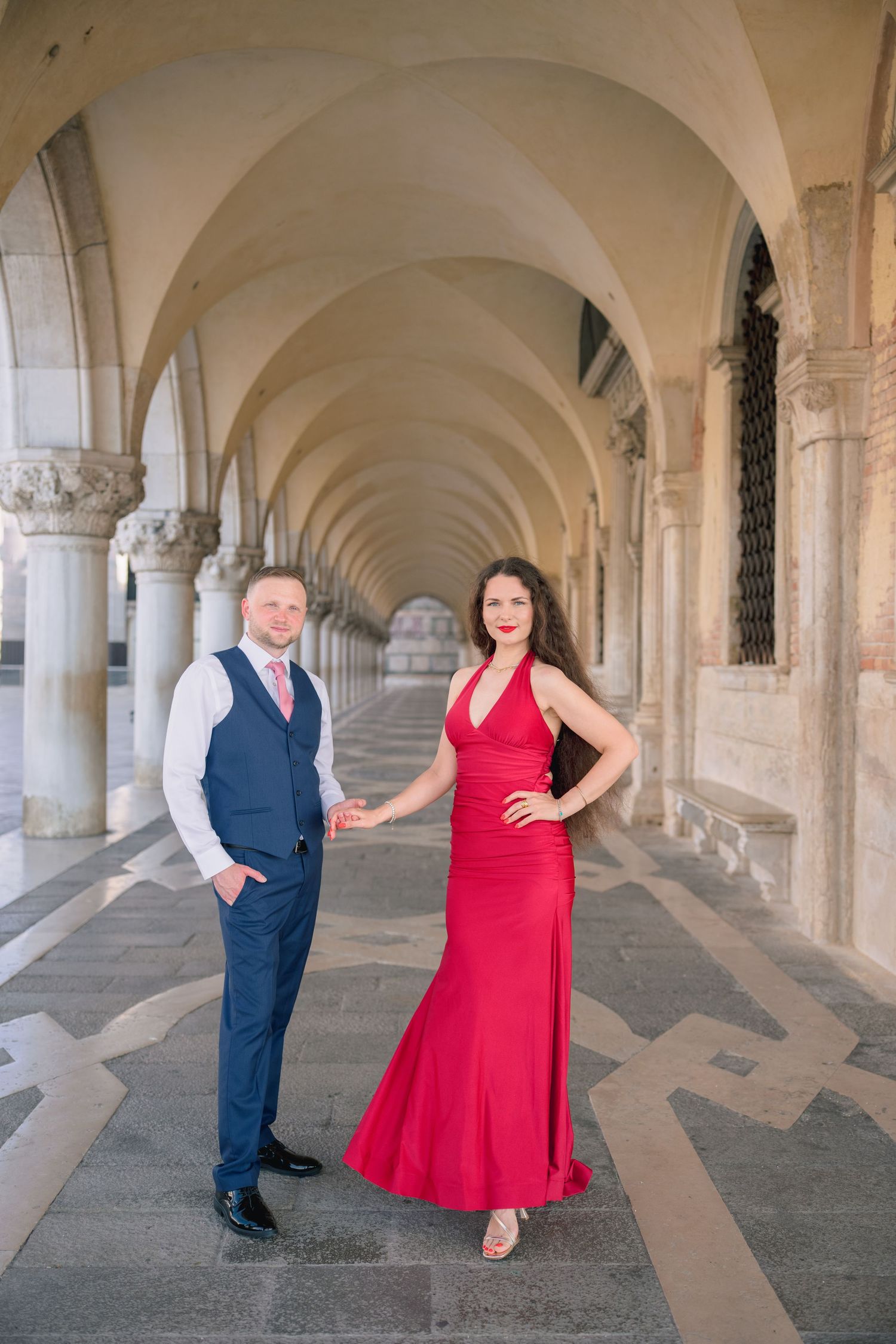 Elegant couple posing under arched colonnade in Venice, woman in red gown and man in blue suit holding hands.