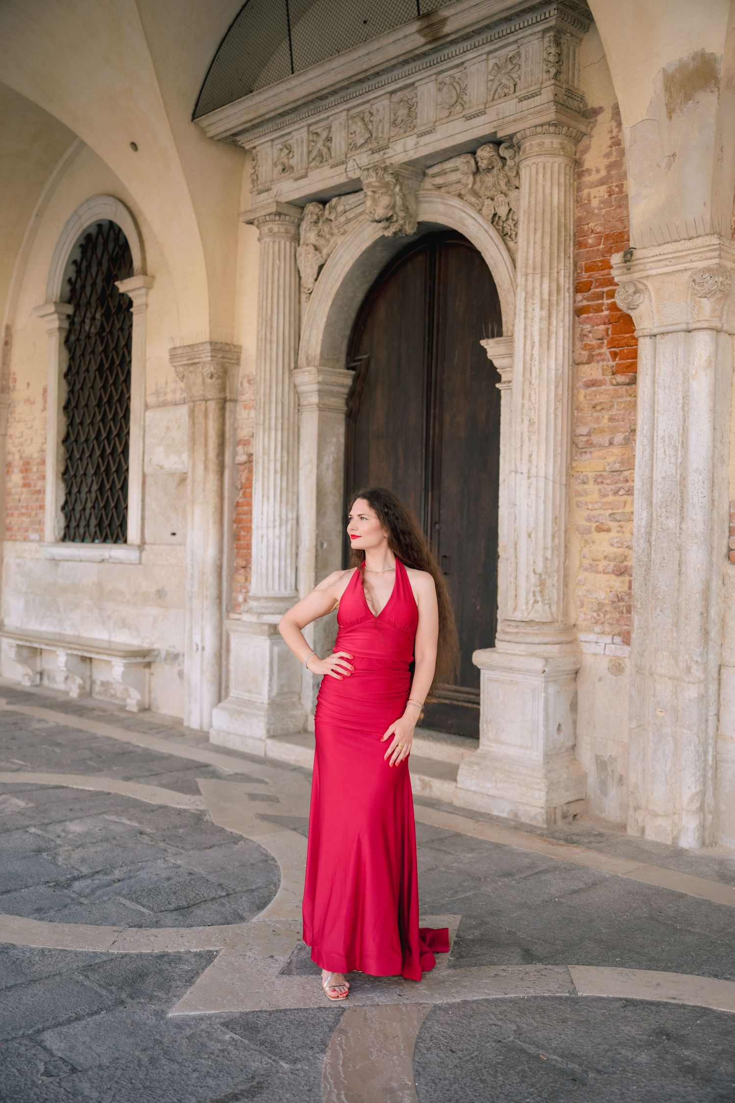 Woman in elegant red gown posing before ornate stone archway and wooden door in historic Venice, Italy.