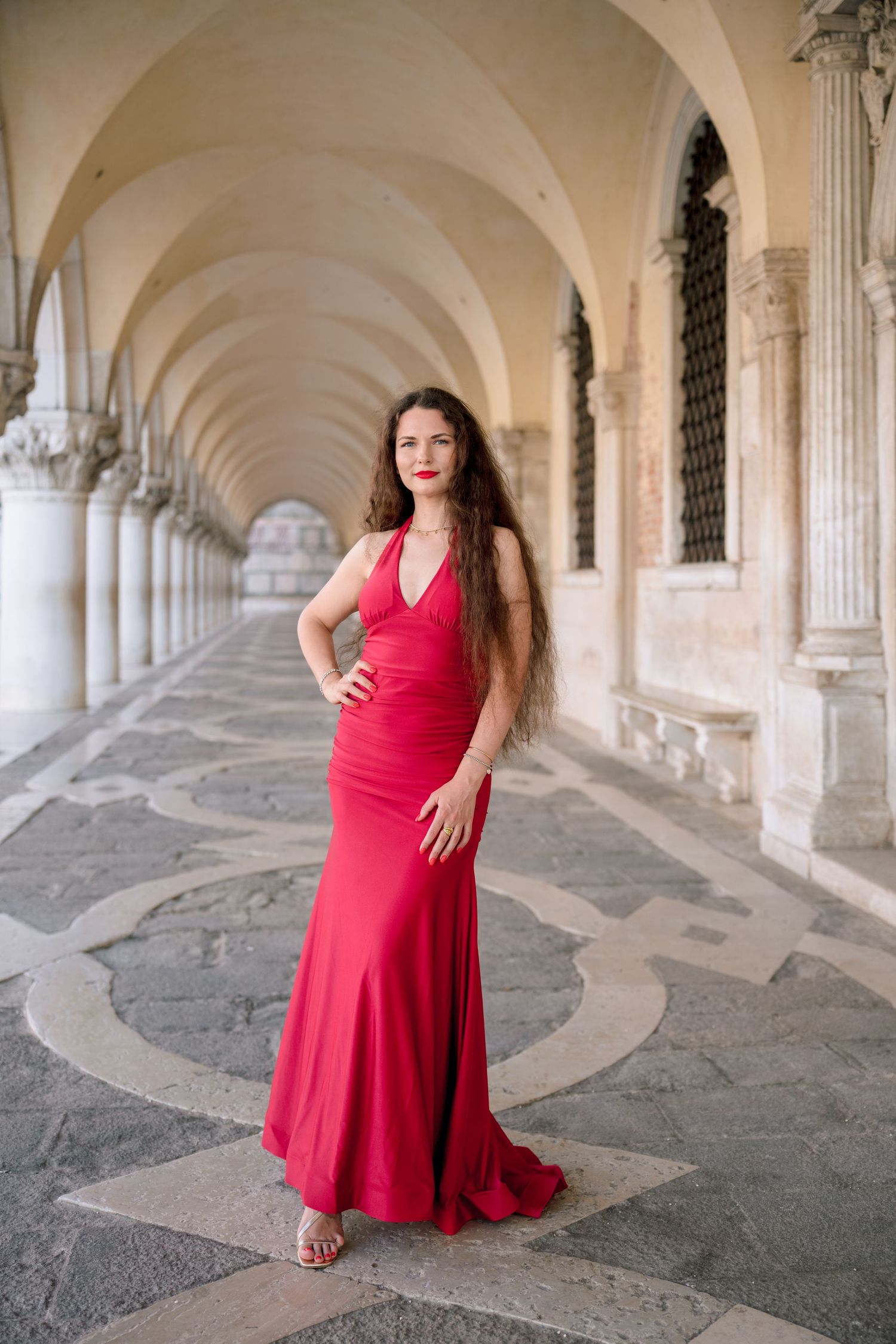 Woman in elegant red gown posing under arched colonnade in Venice, Italy.