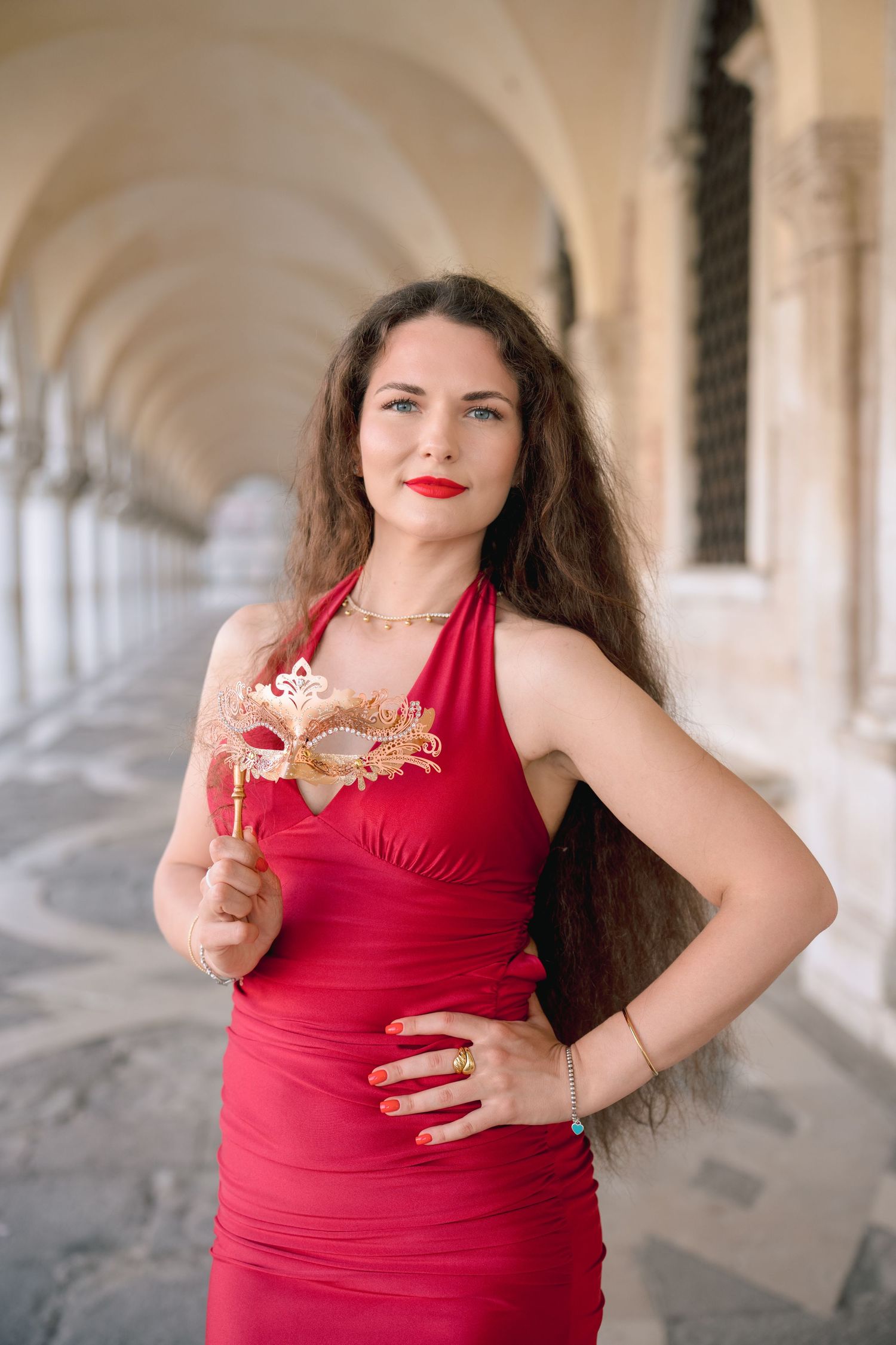 Woman in red dress holding gold Venetian carnival mask under ornate arches in Venice, Italy.