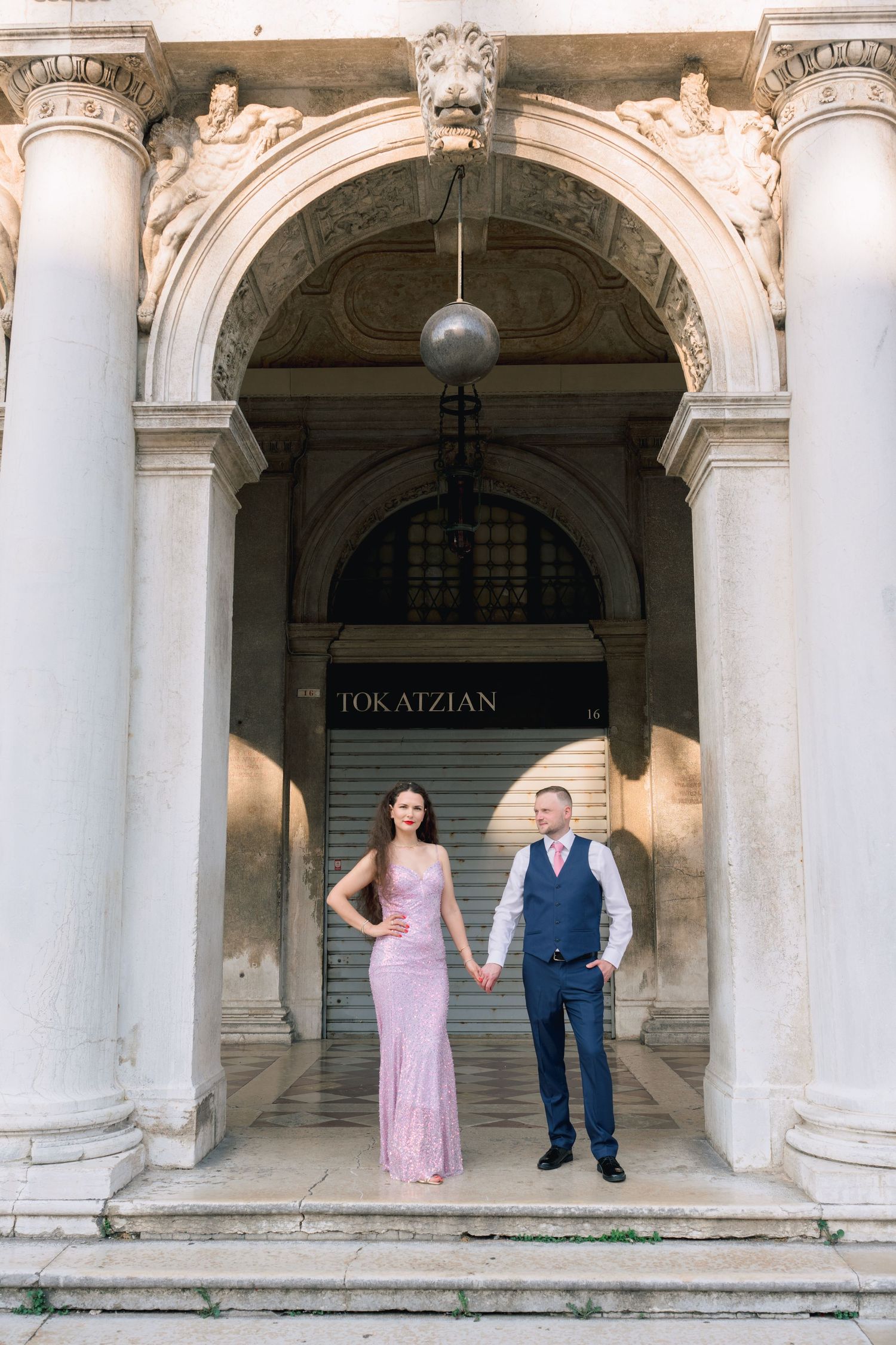 Elegant couple in formal attire holding hands on steps of a classical building with ornate columns and arch.