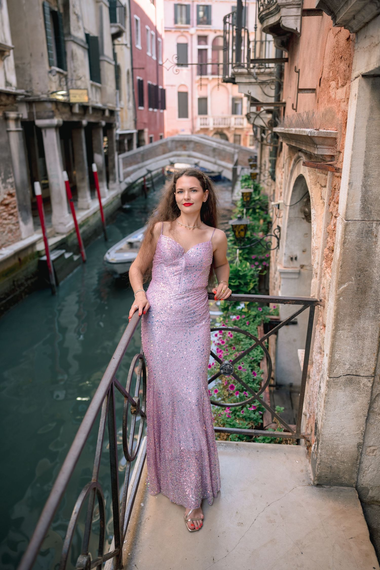 Woman in pink sequin gown poses on Venice canal balcony with bridge and historic buildings in background.