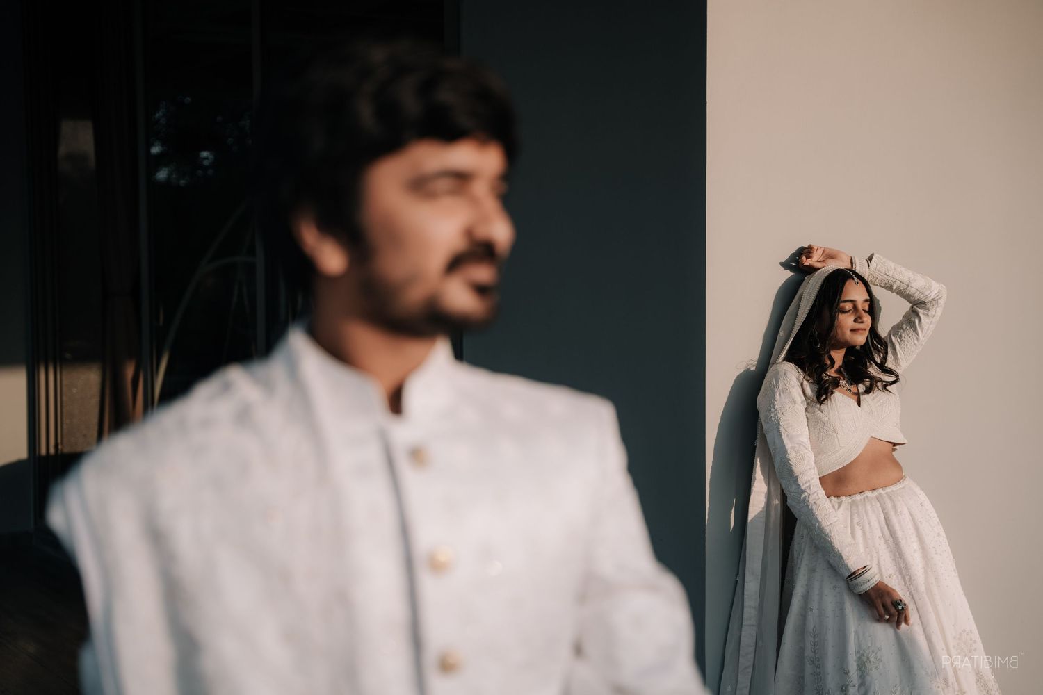 Indian couple in matching white outfits, groom in sherwani foreground, bride adjusting dupatta in soft natural light.