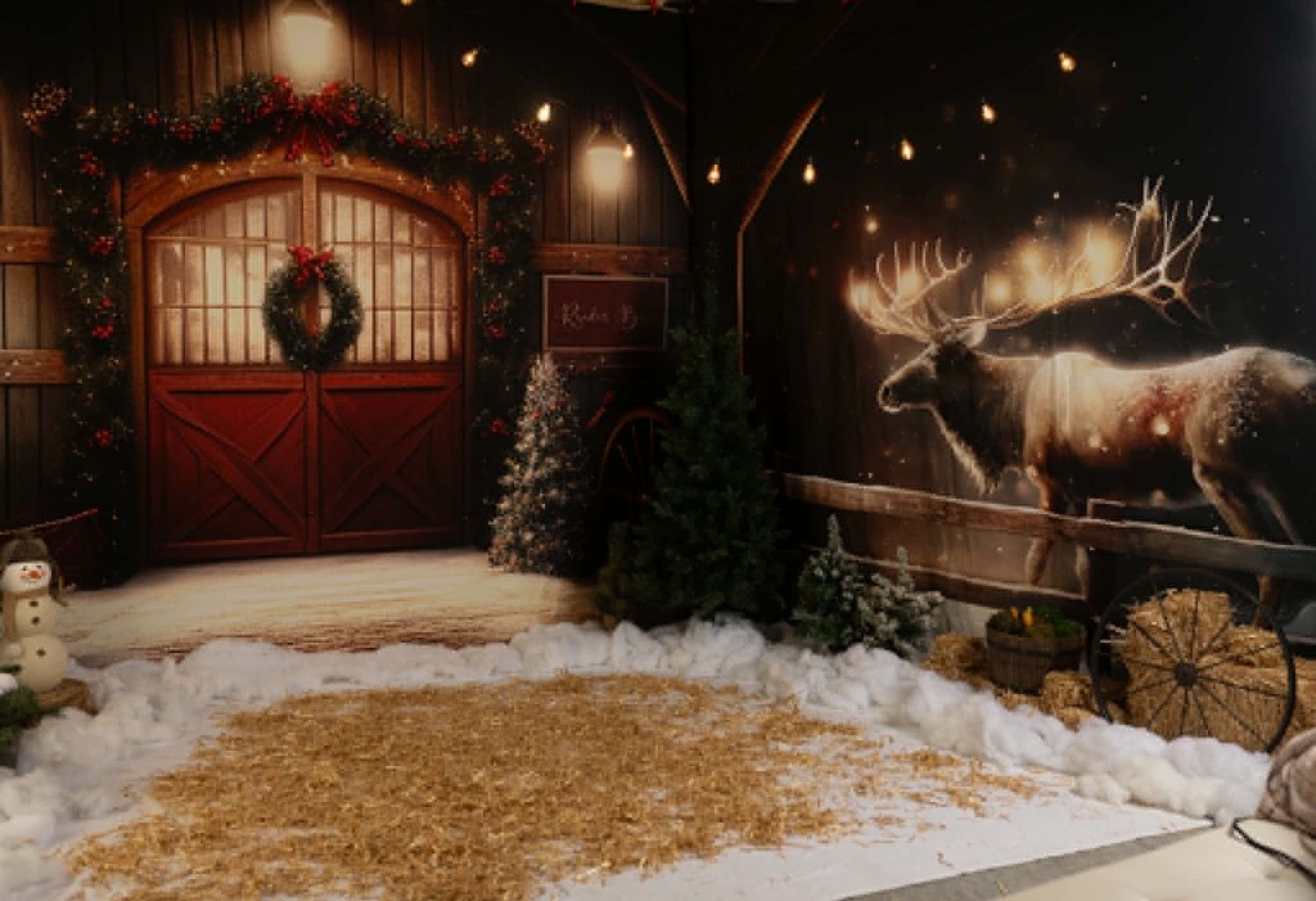 Festive barn scene with Christmas trees, wreath, hay, snow, and a reindeer mural for a holiday photo backdrop.