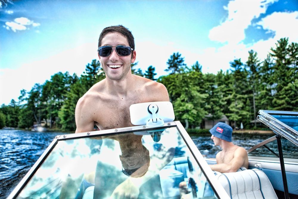 Smiling shirtless man in sunglasses on a motorboat on a lake surrounded by lush green trees.