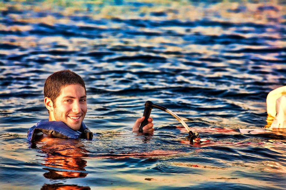 Smiling man swimming in colorful water while holding a camera above the surface.