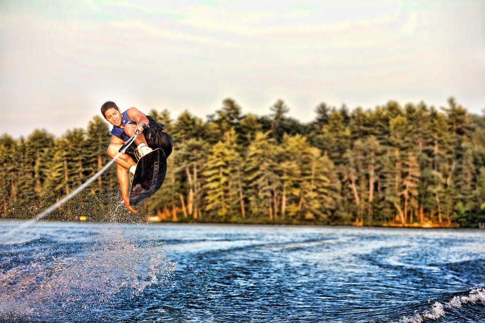 A wakeboarder performs an aerial trick over a lake with lush green trees in the background at sunset.