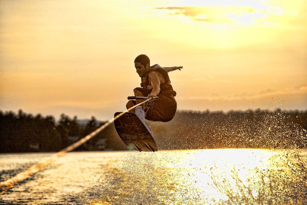 Wakeboarder performing aerial trick over sparkling water at golden sunset, silhouetted against orange sky.