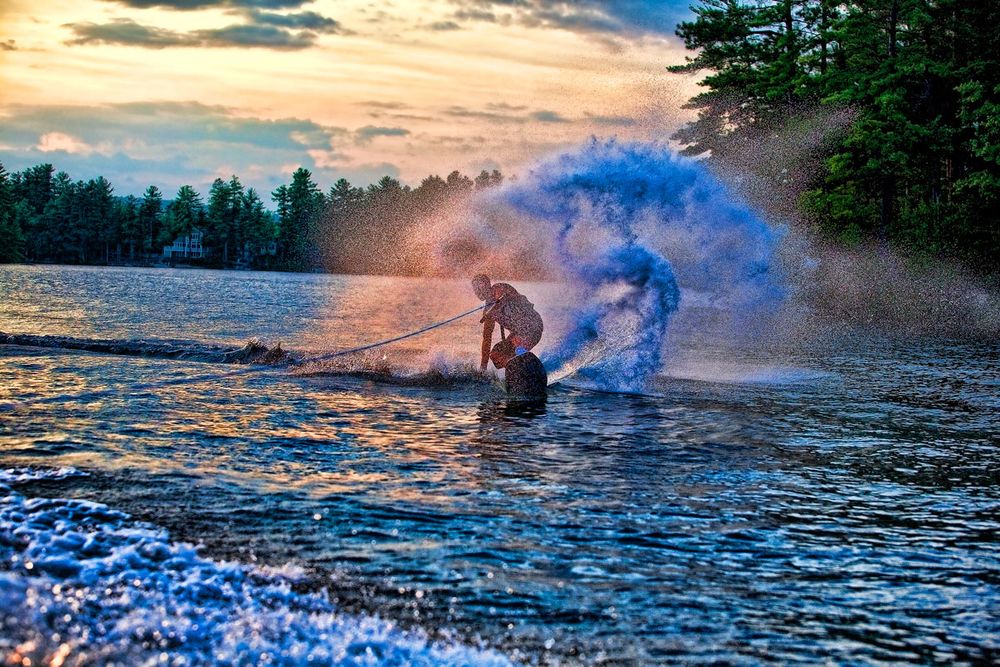 Wakeboarder rides at sunset on a lake with vibrant blue smoke trailing behind, creating a dramatic water sports scene.
