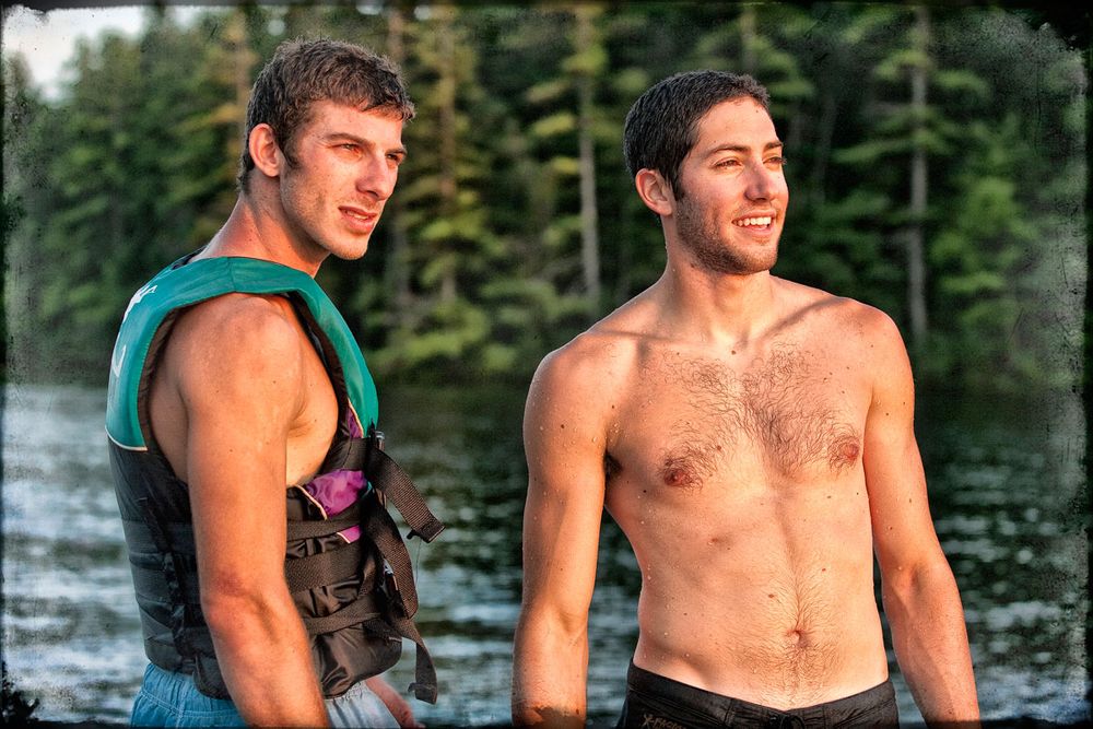 Two young men smiling outdoors near a lake, one wearing a life vest, enjoying a sunny summer day.