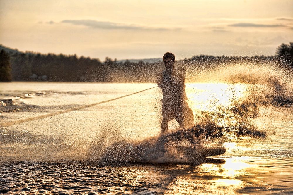 Person water skiing at sunset, creating dramatic splashes on a golden lake.
