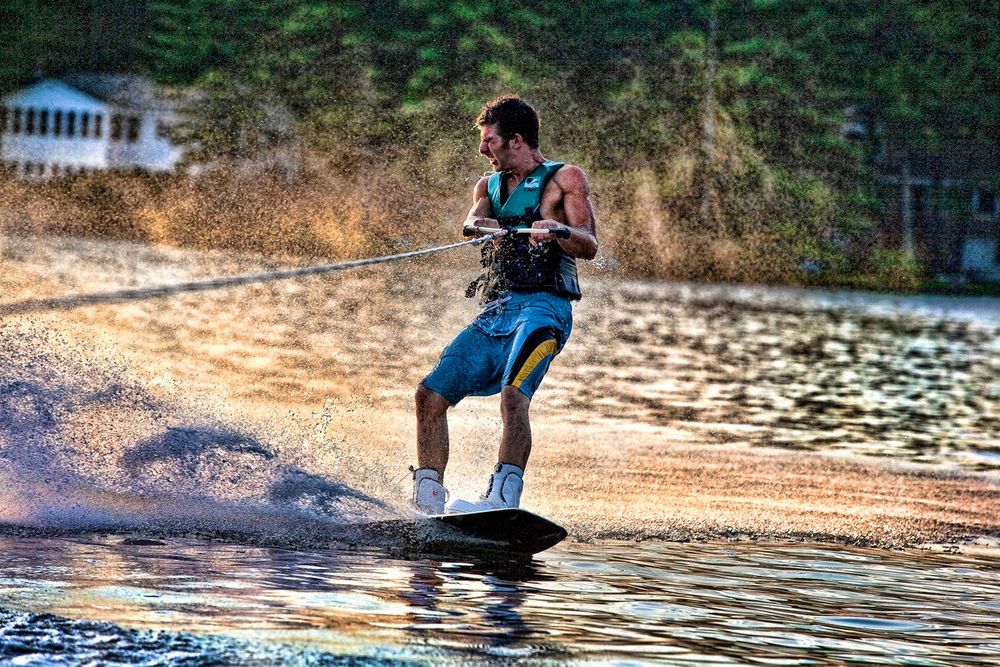 A young man wakeboarding on a lake, creating a dramatic water spray while holding a tow rope at sunset.