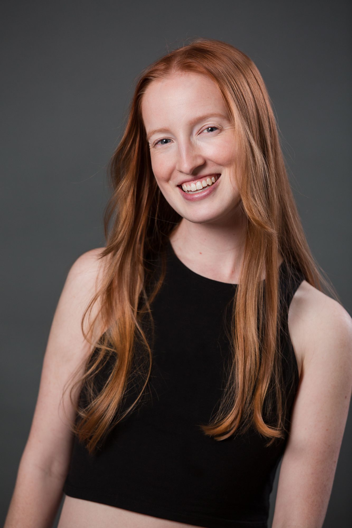 Smiling young woman with long red hair wearing a black crop top, posed against a gray studio background.