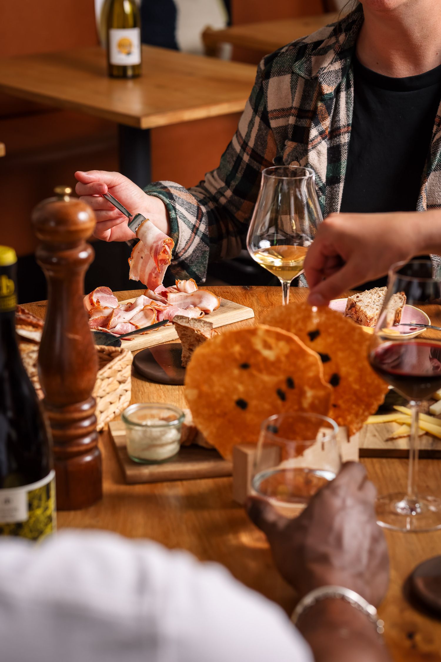 People sharing a charcuterie board with cured meats, cheese, bread, and wine glasses on a wooden table.