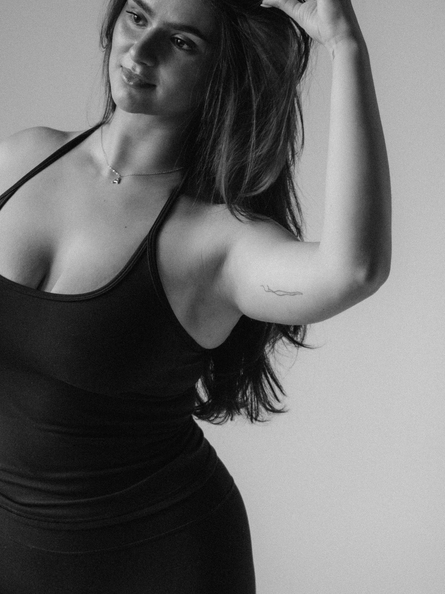 Black and white photo of a young woman in a black dress posing with hand in hair, showing a small arm tattoo.