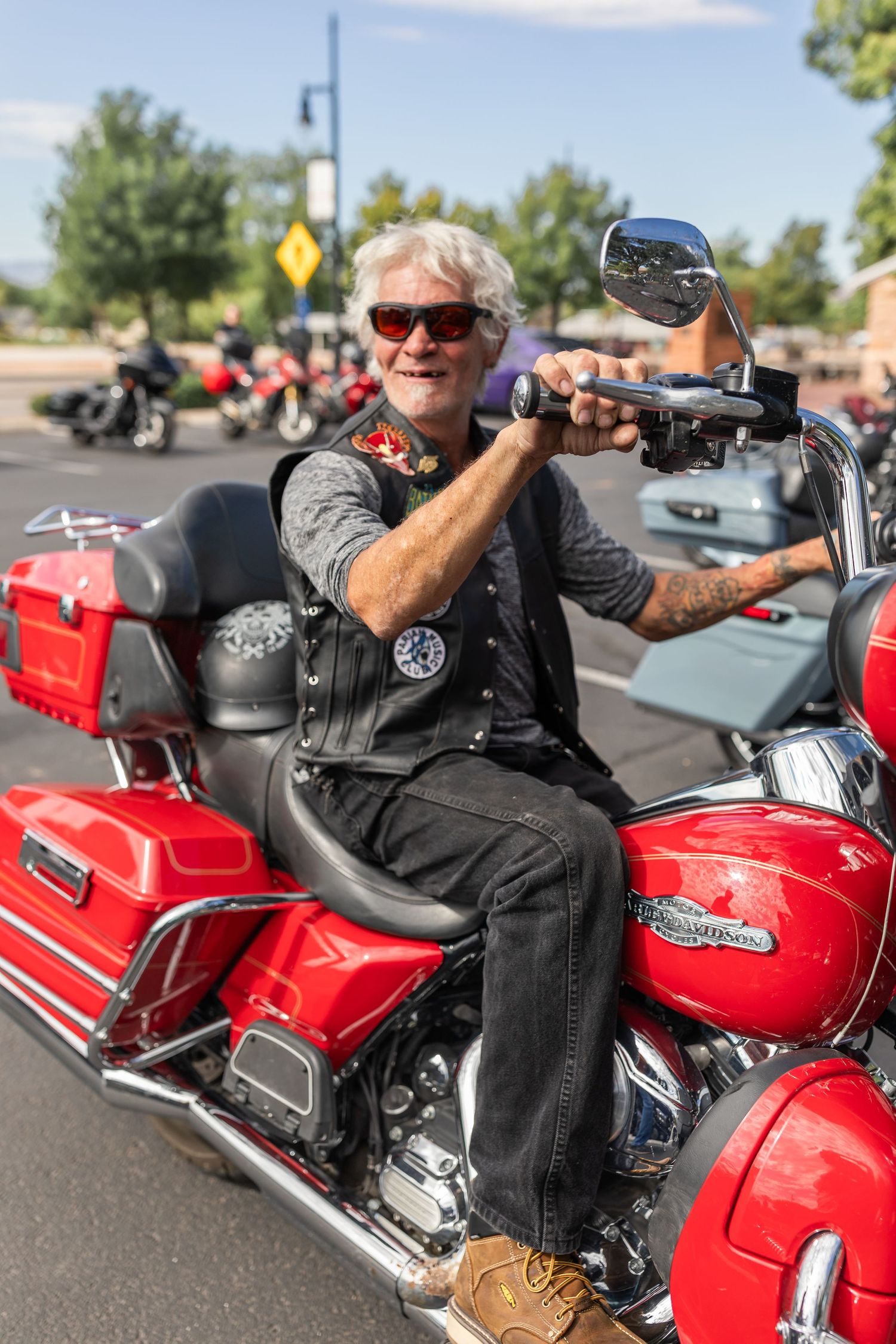 Smiling older man with white hair and sunglasses riding a red Harley-Davidson motorcycle wearing a leather vest with patches.