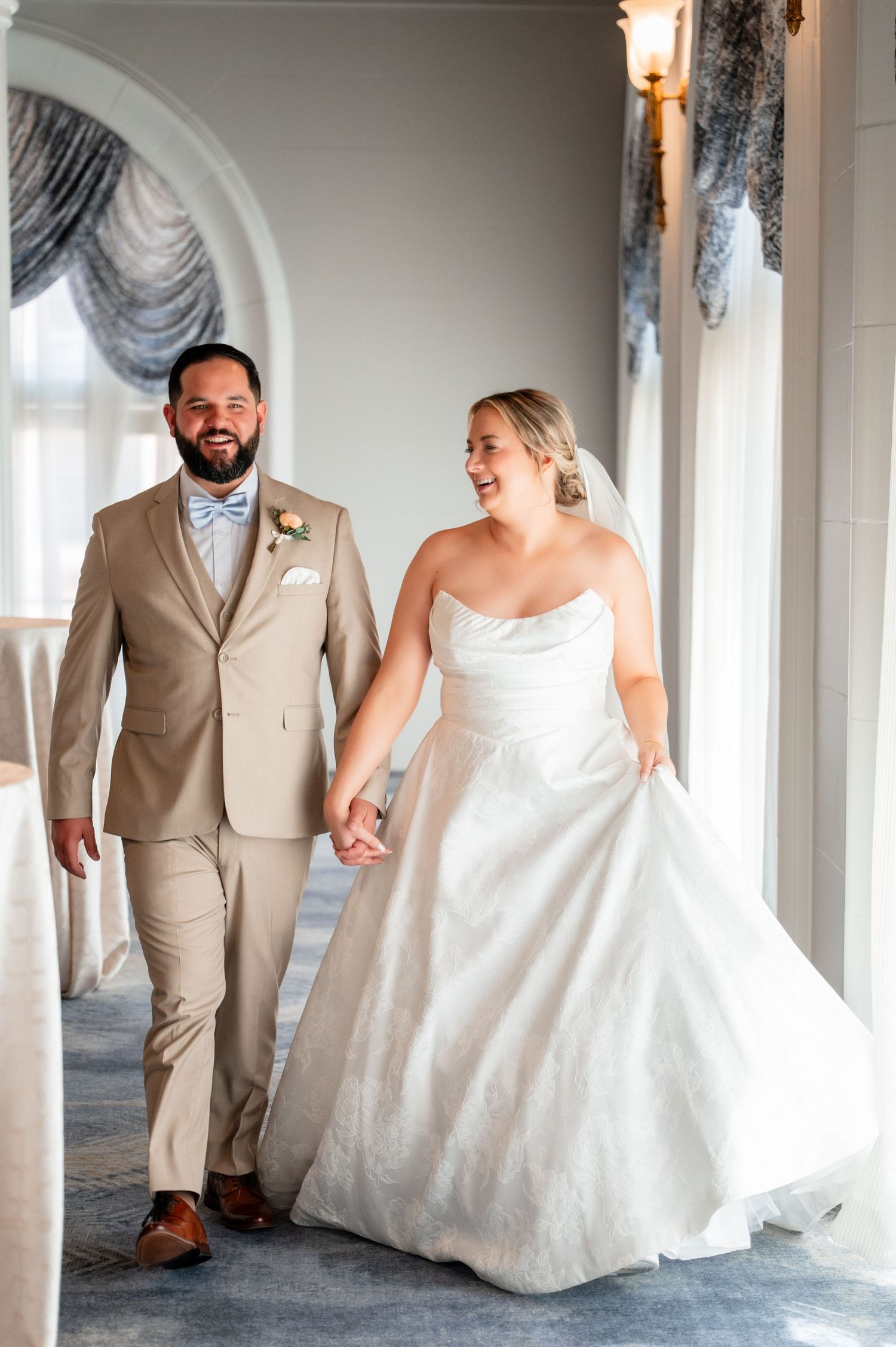 A smiling bride in a white strapless gown holds hands with a groom in a tan suit as they walk together.