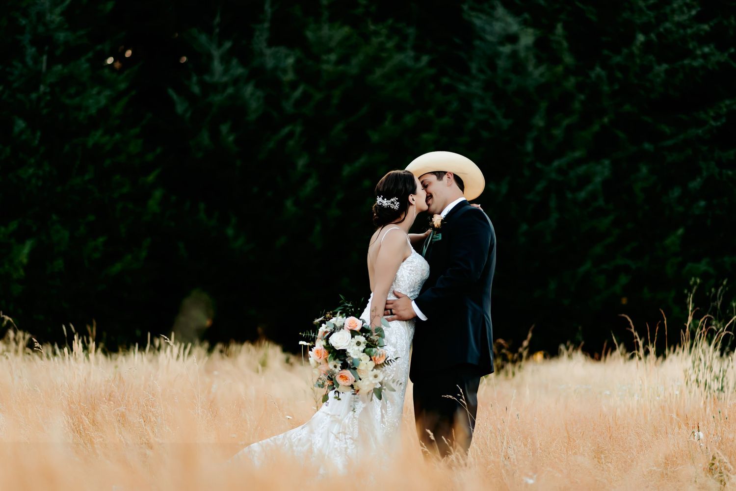 A bride and groom share a romantic kiss in a golden field with lush green trees in the background.