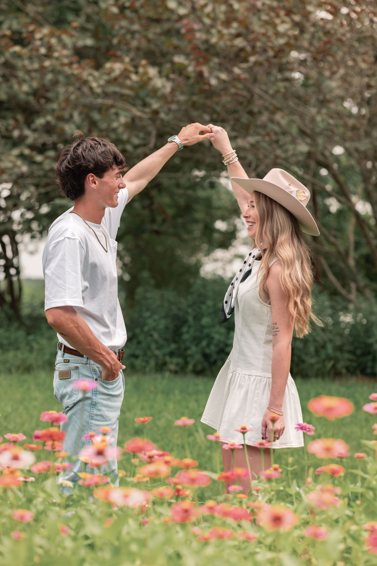 A couple dances joyfully in a flower field, the woman in a white dress and hat, the man in a white shirt and jeans.