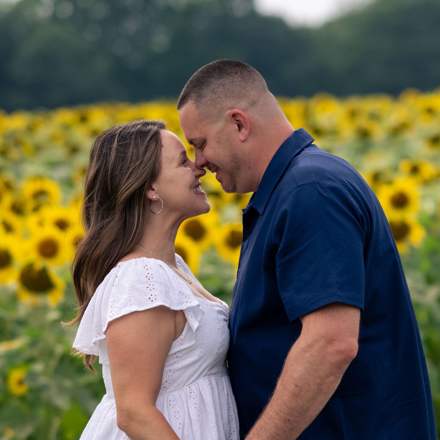 Couple embracing and smiling in a vibrant sunflower field, woman in white top and jeans, man in navy blue shirt.