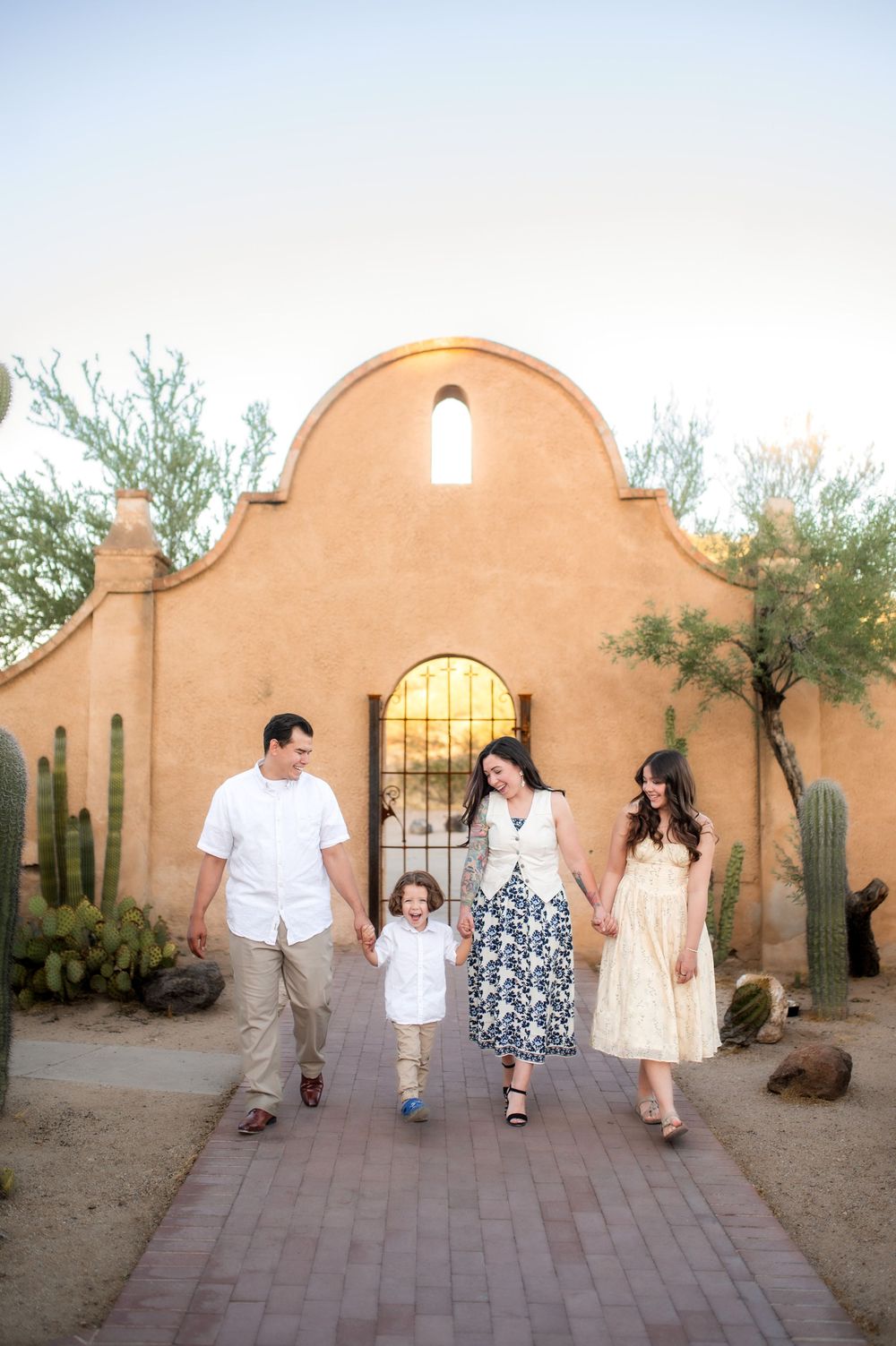 Family of four walking hand-in-hand on a brick path before a adobe-style building surrounded by desert cacti.