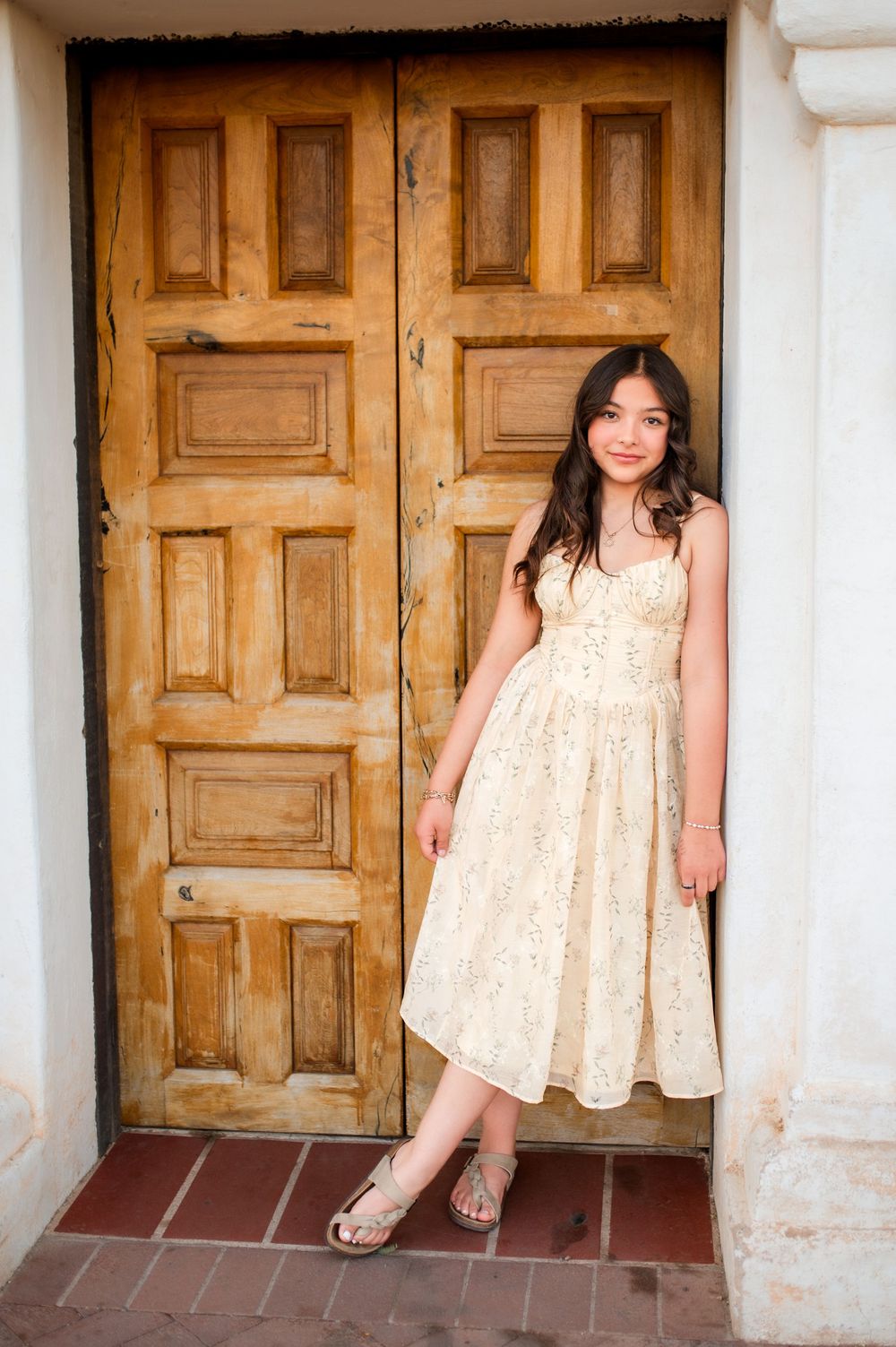 Young girl in cream floral midi dress and sandals posing by rustic wooden double doors.