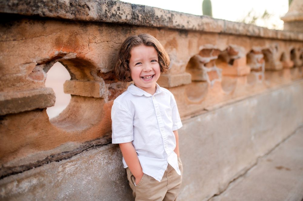 A cheerful young boy with curly hair smiles broadly, leaning against a rustic stone wall in a white shirt.