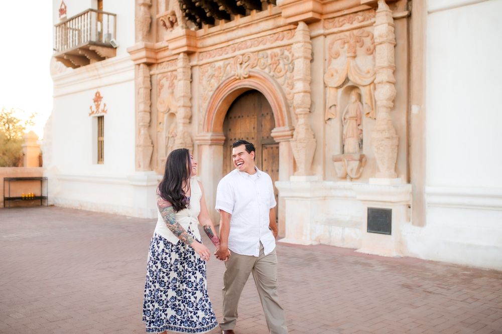 Couple holding hands and smiling at each other in front of an ornate Spanish colonial building facade.