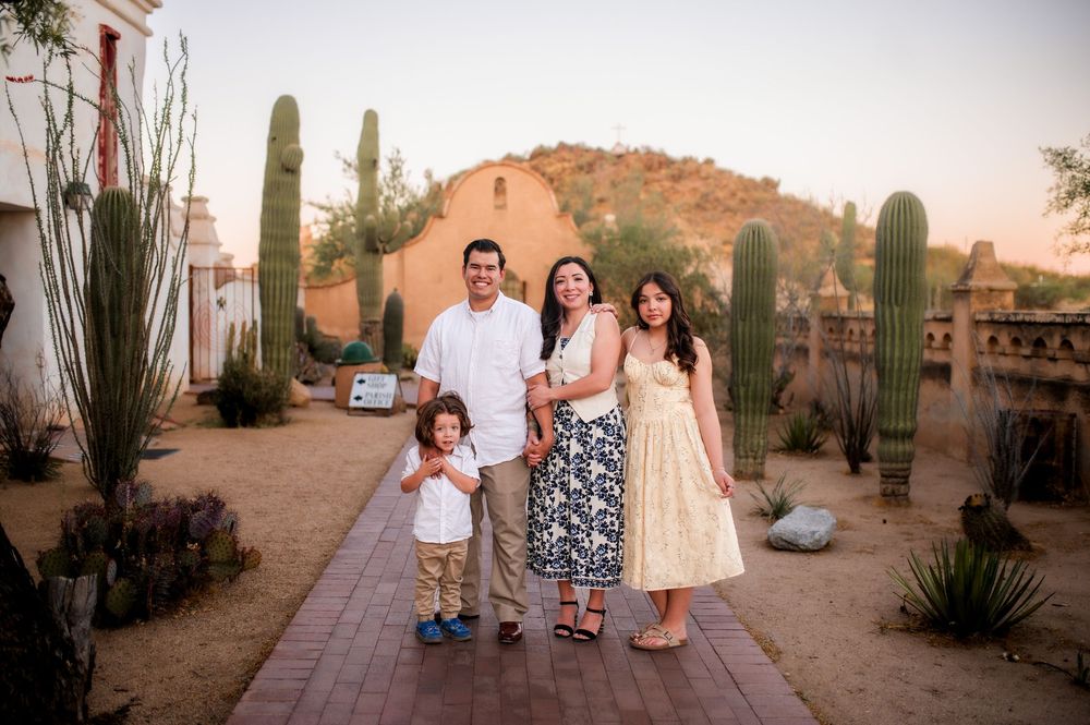 Family of four posing outdoors near saguaro cacti and adobe building in desert landscape at sunset.