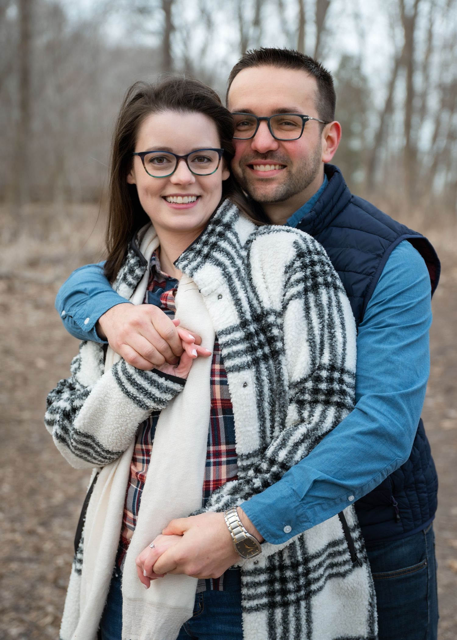 A smiling couple wearing glasses embraces outdoors in winter, the woman in a plaid blanket scarf and the man in a blue vest.