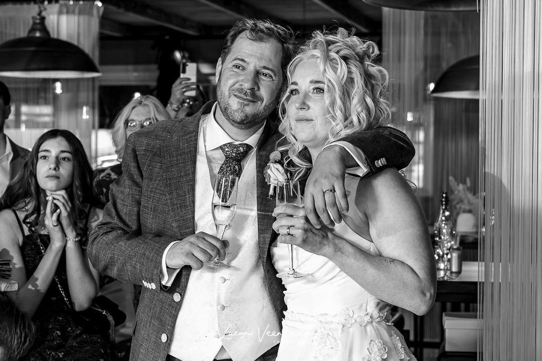 Zwart-wit foto van Vincent en Madelief met hun armen om elkaar heen en champagne in de hand, kijkend naar het filmpje