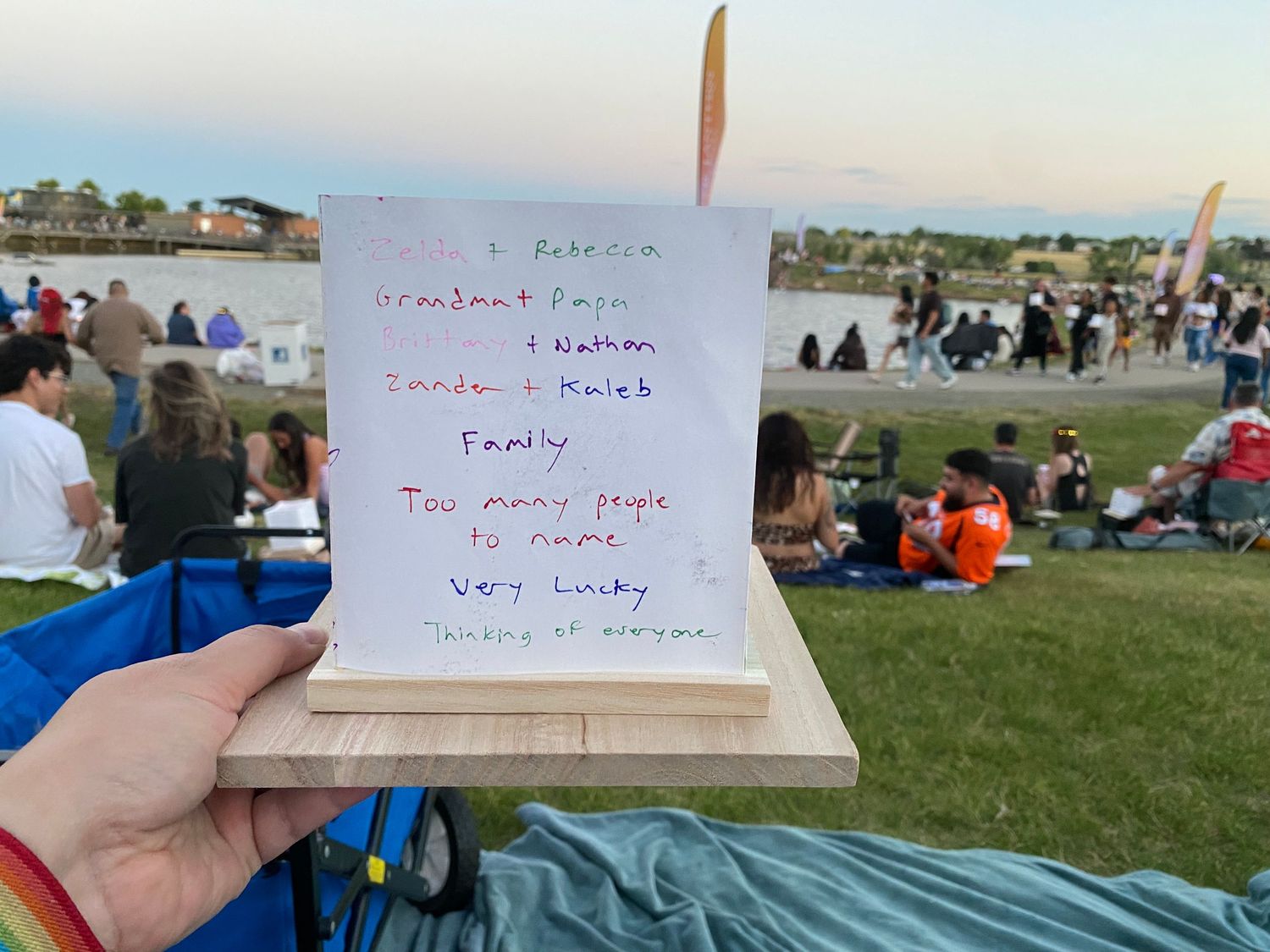 Person holding a handwritten gratitude sign at an outdoor event with crowd and green lawn in background.