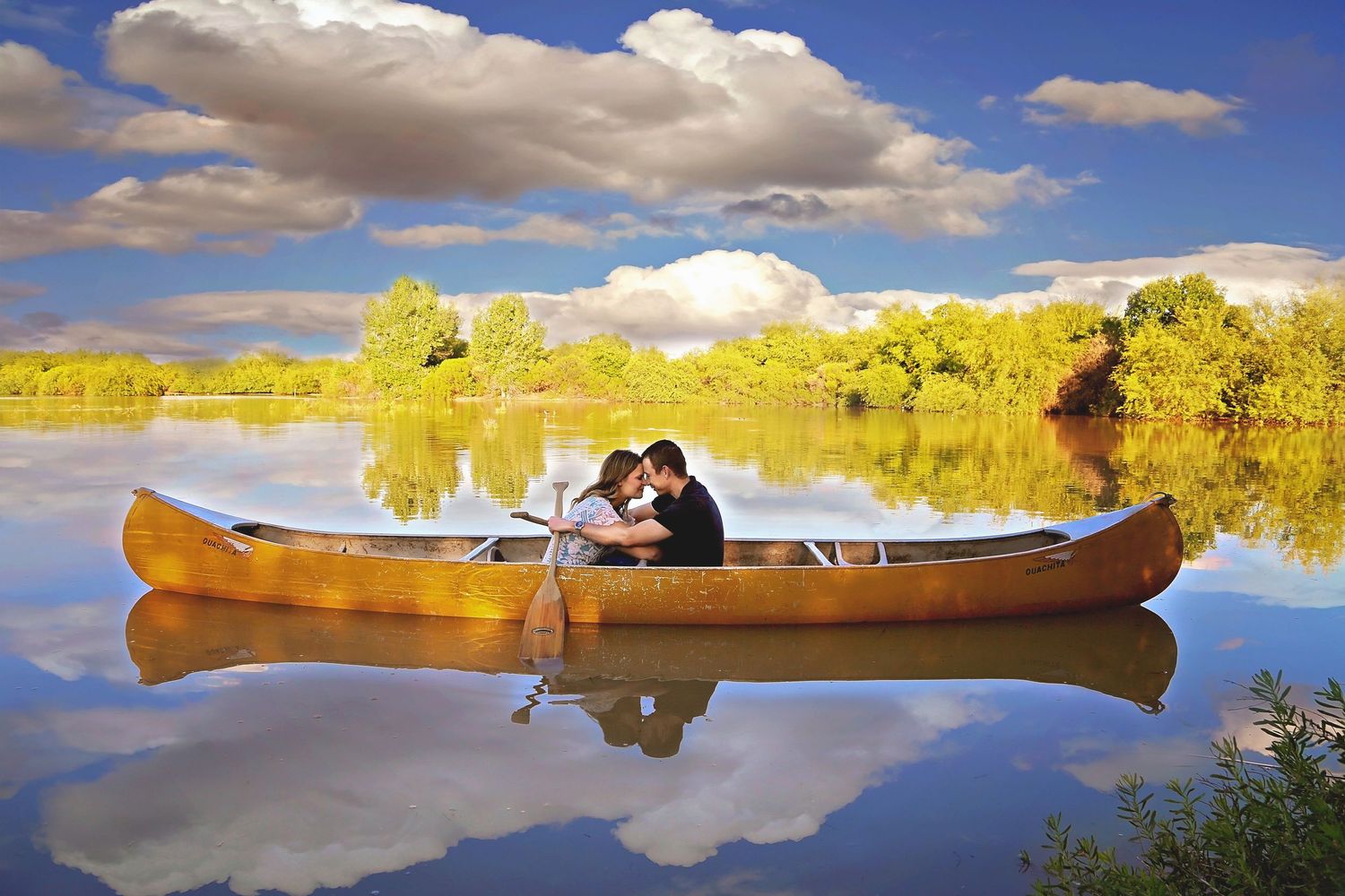 Couple sitting close in a wooden canoe on calm water with golden trees and clouds reflected around them.