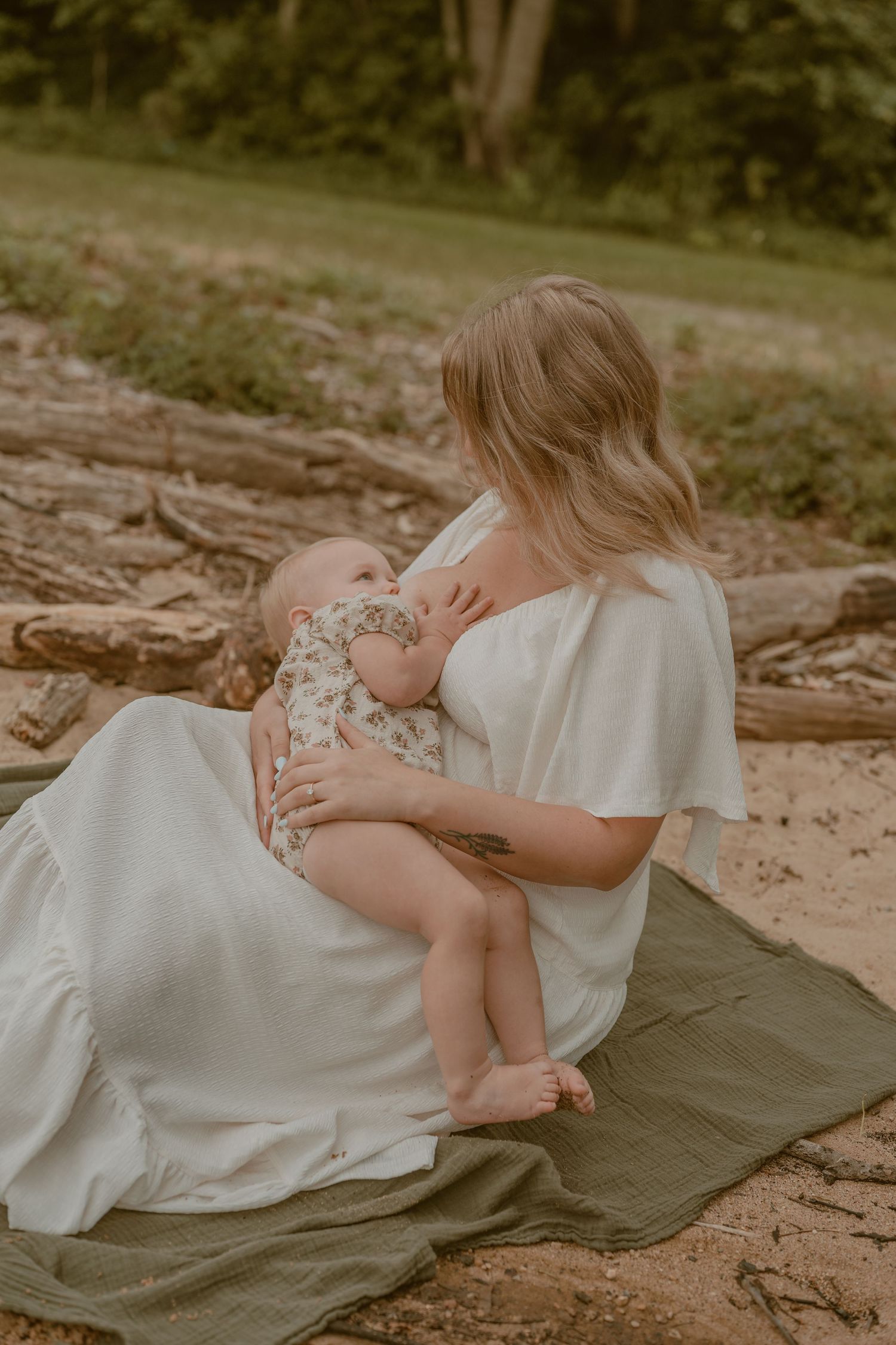 A mother in a white dress breastfeeds her baby outdoors on a green blanket near fallen logs.