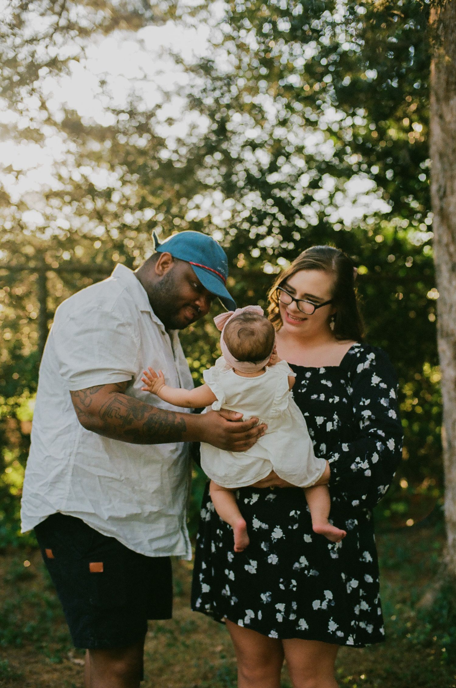 A couple stands outdoors holding their baby in golden sunlight, surrounded by lush green trees.