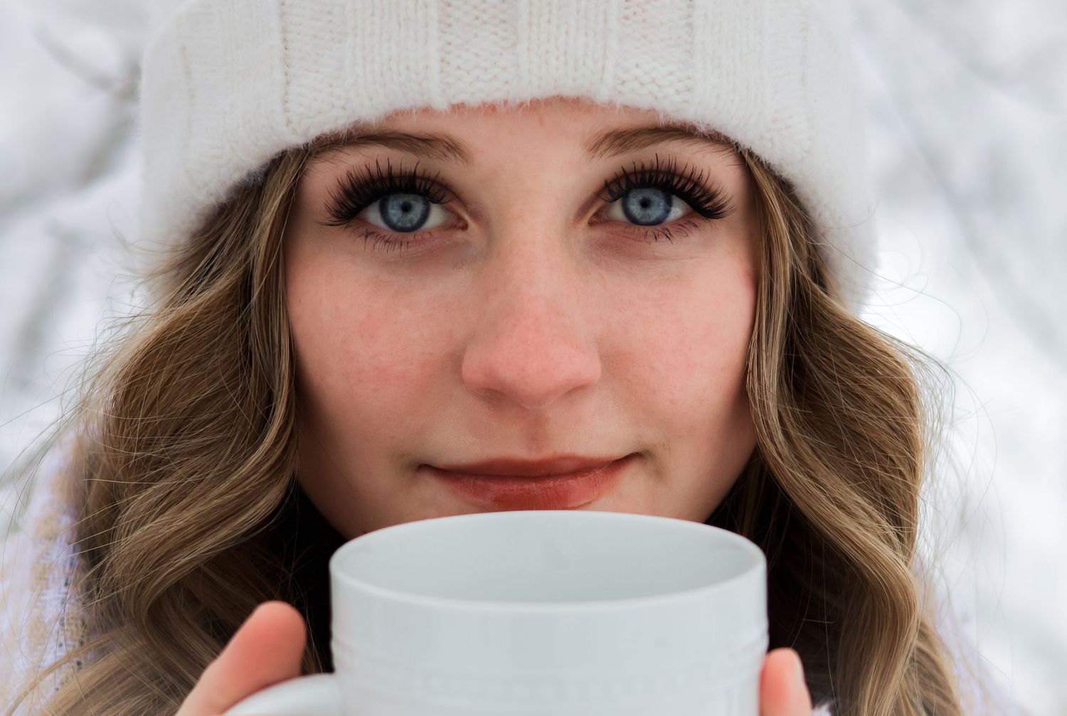 Close-up portrait of blue-eyed woman in white winter hat holding a white mug outdoors.