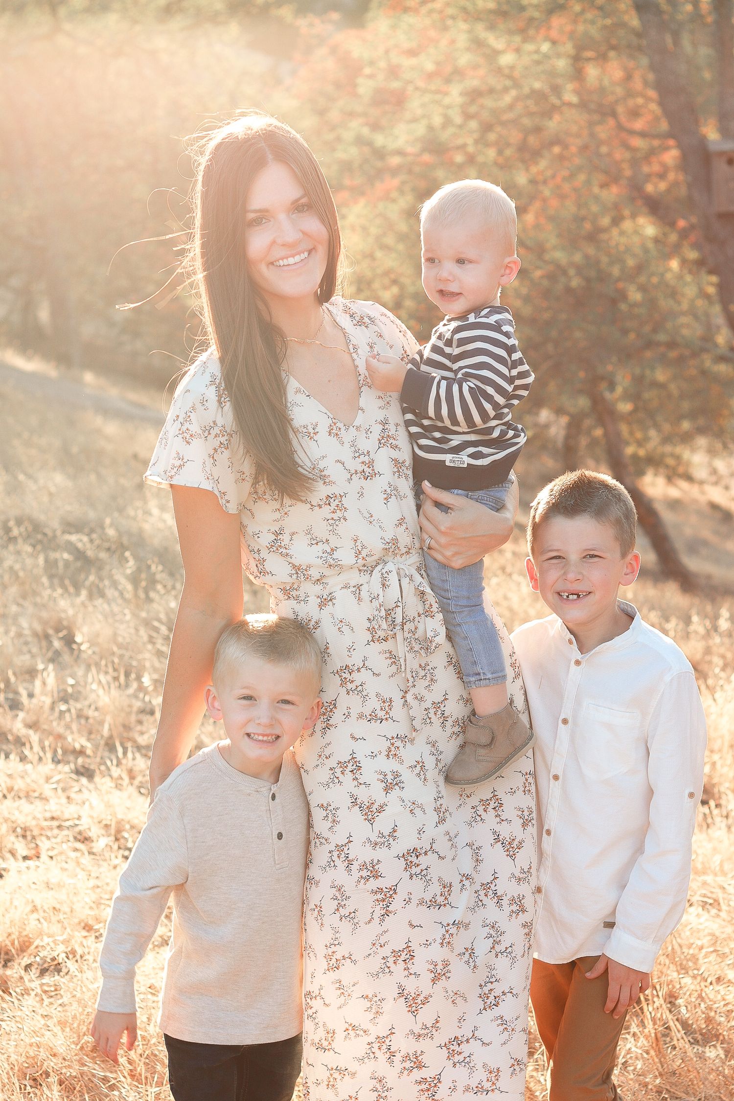 Smiling mother in floral dress holding baby while two young boys stand beside her in sunny autumn field.
