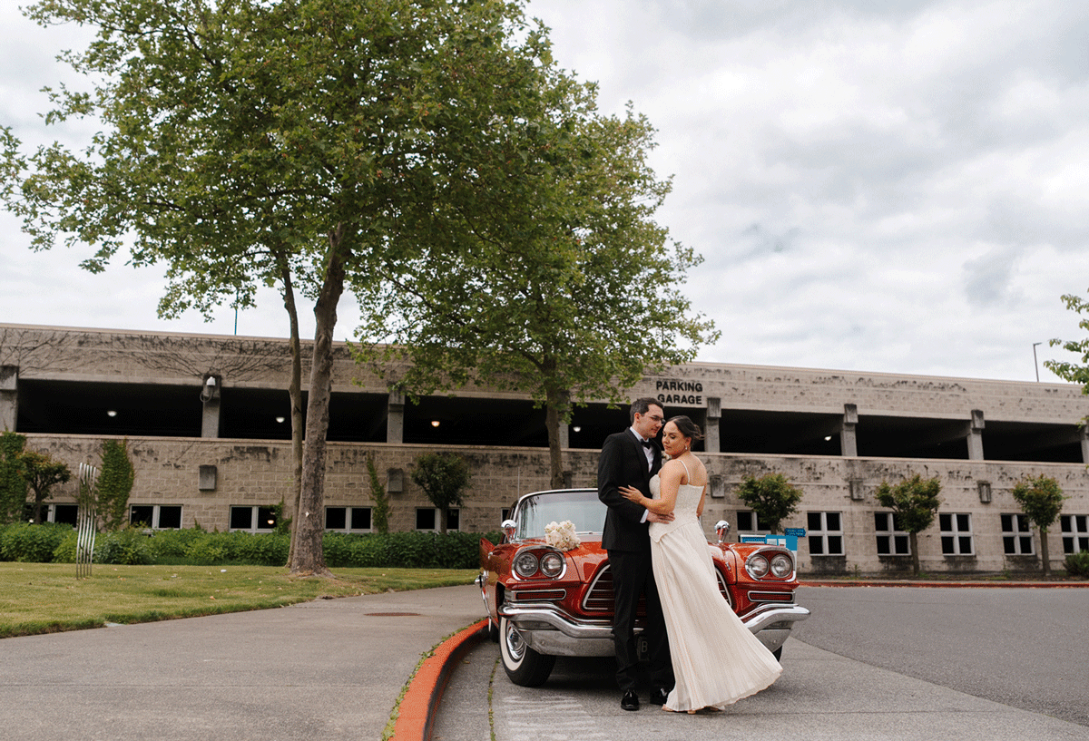 Bride and groom share a romantic kiss beside a classic red car outside a stone building on their wedding day.