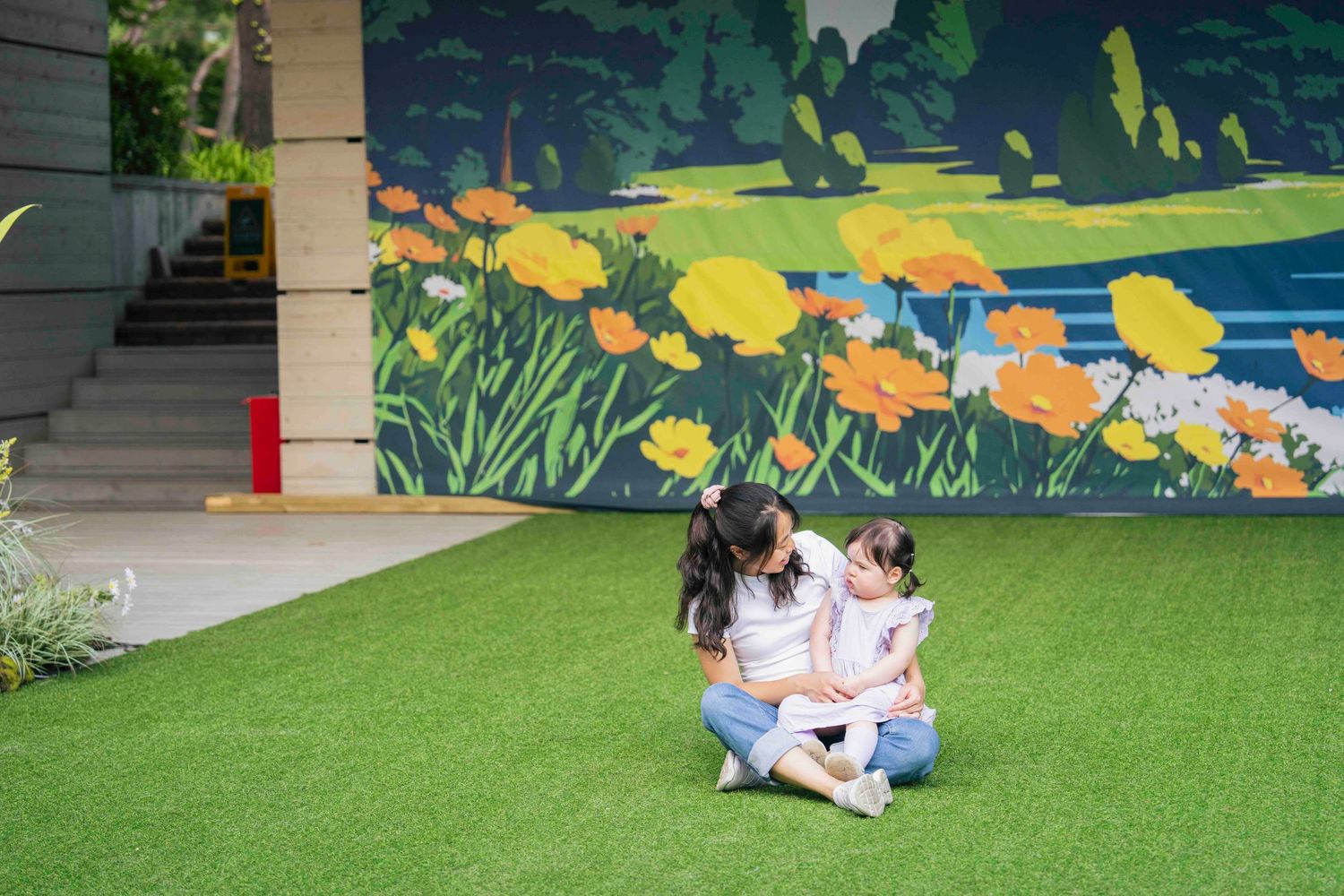 Mother and daughter sitting on artificial grass in front of a colorful floral mural outdoors.