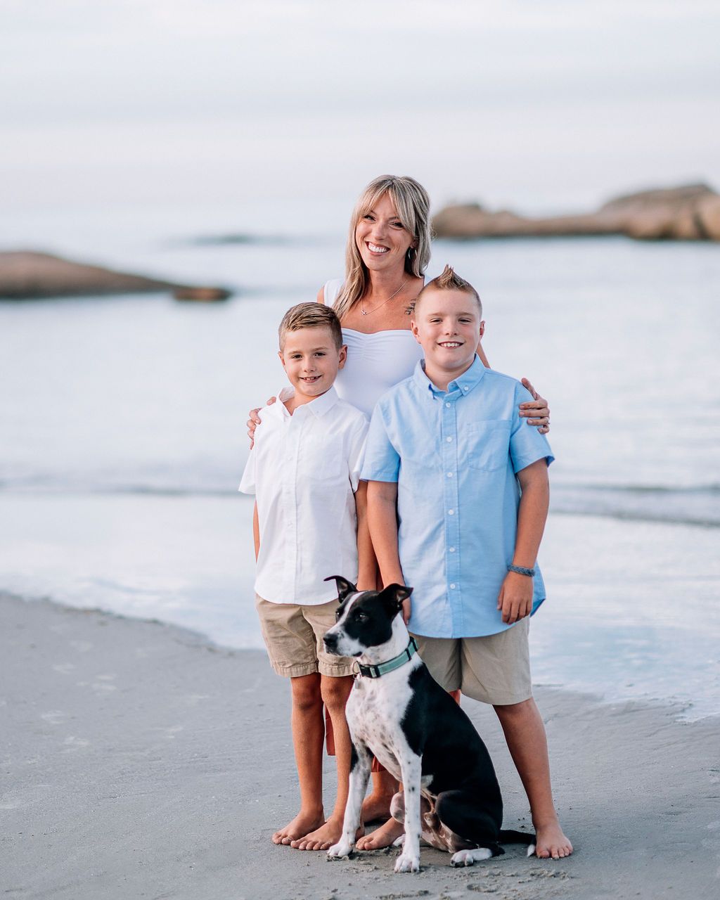 A woman and two boys pose with a black and white dog on a sandy beach at sunset.