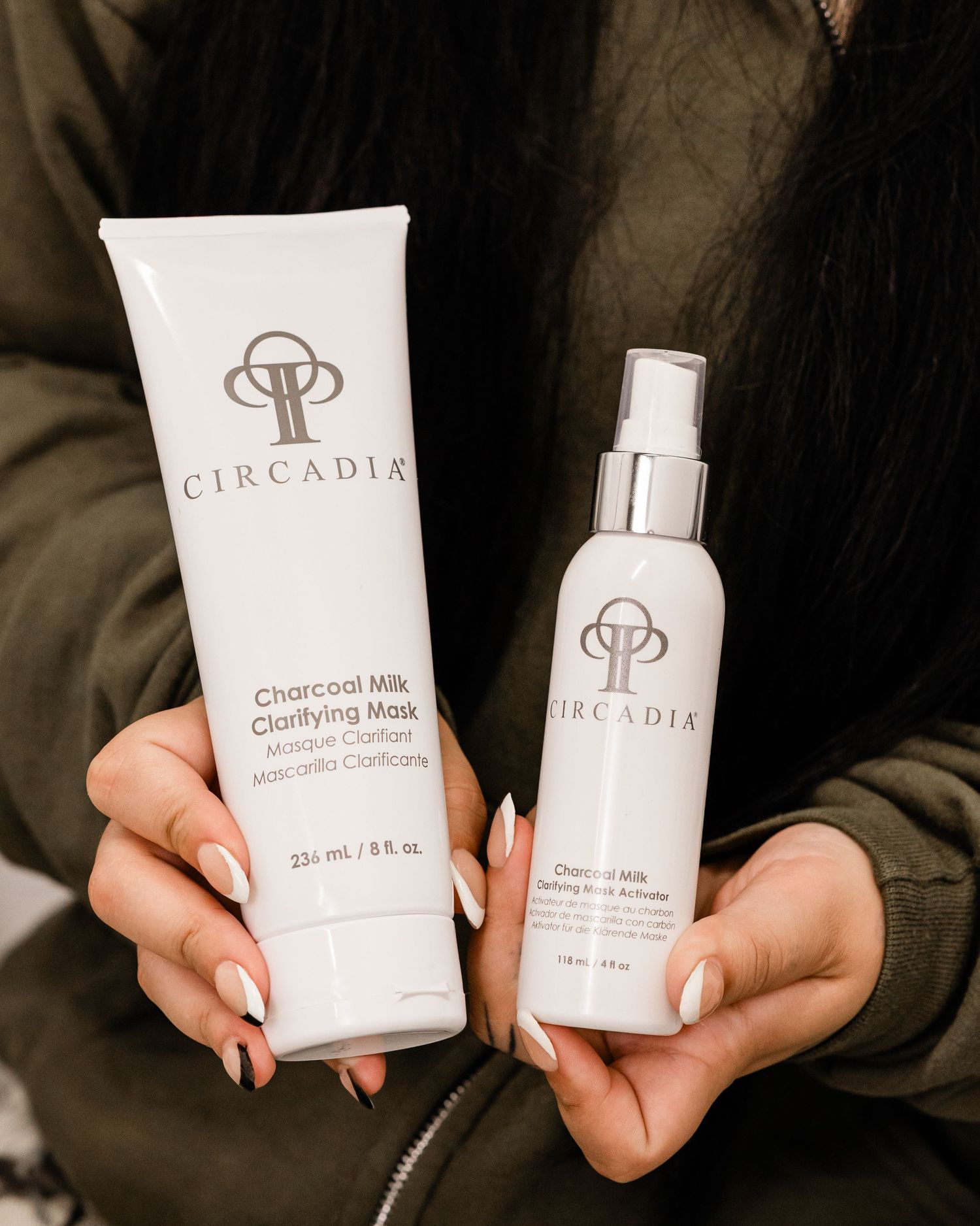 Hands holding Circadia Charcoal Milk Clarifying Mask tube and spray bottle against a dark jacket background.