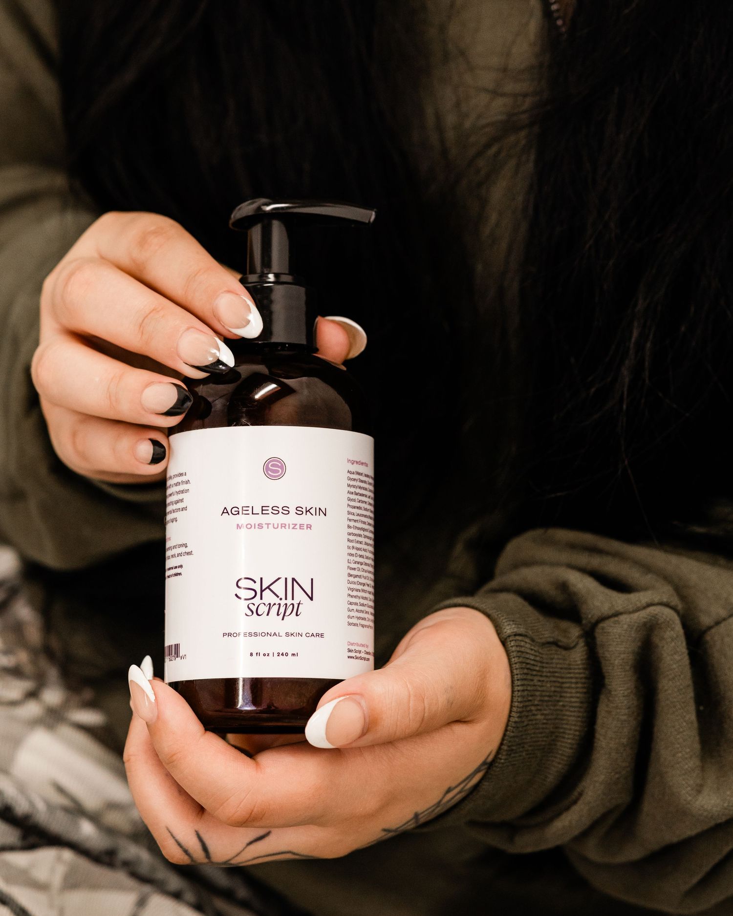 Hands holding Skin Script Ageless Skin Moisturizer in a dark amber pump bottle.