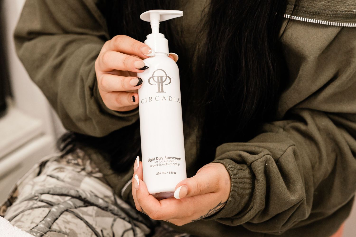 Woman holding a white pump bottle of Arcadia skincare lotion while wearing a green hoodie.